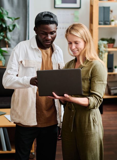 Two young intercultural employees in casualwear looking at screen of laptop Two young intercultural employees in casualwear looking at screen of laptop held by blond businesswoman during discussion